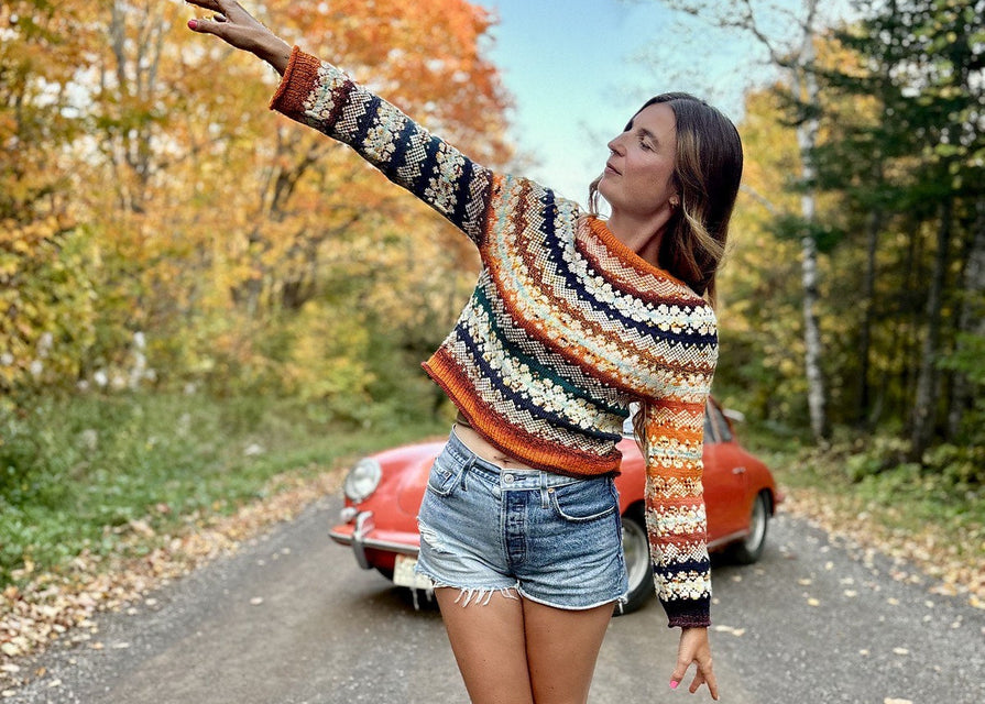 Woman in a colorful sweater and denim shorts standing on a road with autumn trees and a vintage car in the background.
